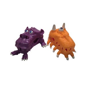 May include: Two whimsical toy figures. One is a purple hippo-like creature with large eyes and small feet. The other is an orange, spiky, monster-like figure with white horns and eyes. Both are set against a white background.