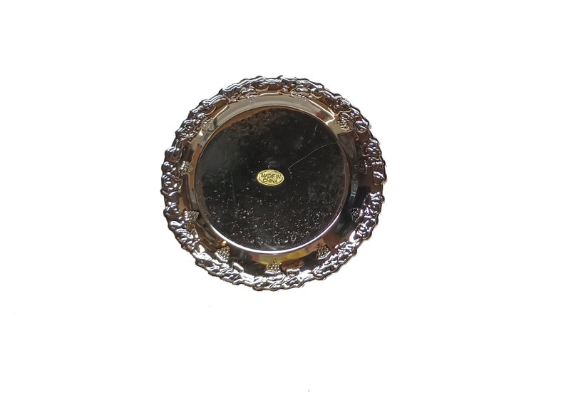 May include: Silver-colored round serving tray with a decorative embossed border. The tray has a label that reads "Made in China".