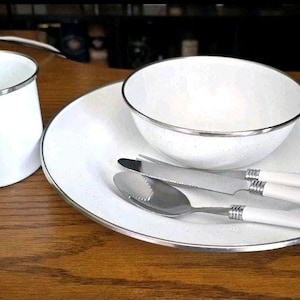 Peut inclure: Un ensemble de vaisselle en émail blanc avec une finition mouchetée et des bords argentés. Le set comprend une tasse, un bol, une assiette, un couteau et une cuillère. Le couteau et la cuillère ont des manches blancs et des têtes en acier.