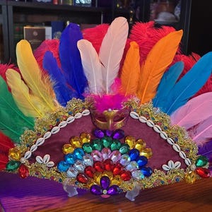 May include: A decorative fan featuring a vibrant array of multi-colored feathers in shades of red, yellow, blue, orange, and purple. The fan is adorned with gold trim, white cowrie shells, and a central arrangement of colorful faceted gems.