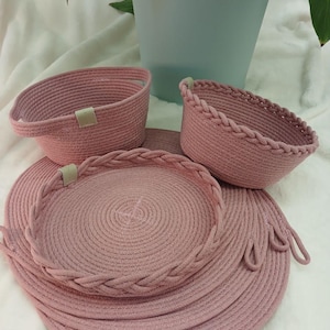 May include: A set of pink rope baskets and placemats. The set includes two oval baskets, a round tray, and two placemats. The baskets have a braided trim and a small tag. The placemats and tray are made of coiled rope.