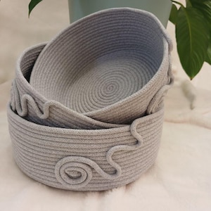 Peut inclure: Trois paniers en corde grise de tailles différentes sont imbriqués. Les paniers présentent un motif en spirale sur le fond et des volutes décoratives sur les côtés. Fabriqués en matériau tissé doux.