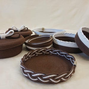 May include: A collection of handcrafted brown and white rope baskets and containers. Some have lids with white bow accents, while others feature braided or striped edges. The various sizes and shapes offer versatile storage solutions.