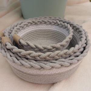 May include: Set of three gray and white rope baskets. The baskets are nested, with a braided rope design around the rim. The interior of the baskets is white, and the exterior is gray. The baskets have small leather accents.