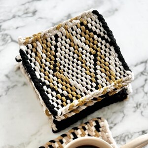 May include: Set of four woven coasters in black, white, and gold. The coasters are square and have a textured surface. A coffee mug is sitting on one of the coasters.