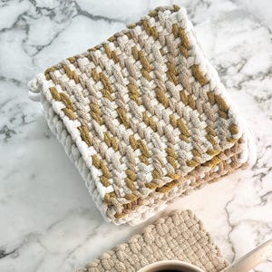 May include: Two woven cotton pot holders in beige and mustard yellow. One pot holder is being used as a coaster for a white ceramic mug filled with black coffee.