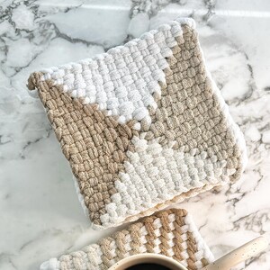 May include: Two woven cotton pot holders in beige and white. The pot holders are square shaped and have a geometric pattern. A white mug with black coffee is sitting on top of one of the pot holders.