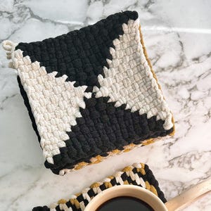 May include: Two woven pot holders, one black and white and one with black, white, and gold stripes. A white mug with a black handle sits on the gold striped pot holder, filled with black coffee.