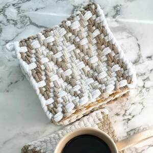 May include: Two woven potholders in white, tan, and brown. One potholder has a small ceramic mug with dark liquid. The potholders are square and have a woven design. The mug has a handle and is filled with a dark liquid.