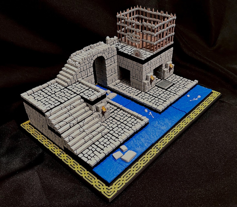 Puede incluir: Diorama en miniatura de un castillo de piedra con un r&iacute;o azul, escaleras, un arco y una estructura en forma de jaula en la parte superior. La base tiene un borde dorado. Modelo para juegos de mesa o exhibici&oacute;n.