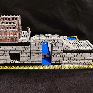 Puede incluir: Un modelo de castillo en miniatura con muros de ladrillo gris, una puerta arqueada azul y una estructura de jaula de madera en la parte superior. La base es negra con un patr&oacute;n geom&eacute;trico dorado. El modelo es para juegos de mesa.