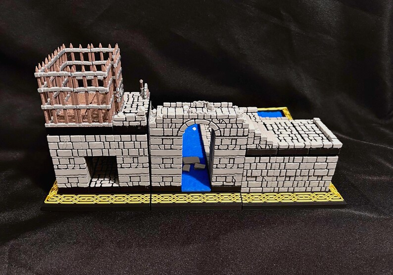 Puede incluir: Un modelo de castillo en miniatura con muros de ladrillo gris, una puerta arqueada azul y una estructura de jaula de madera en la parte superior. La base es negra con un patr&oacute;n geom&eacute;trico dorado. El modelo es para juegos de mesa.