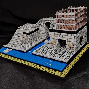 Puede incluir: Diorama en miniatura de un castillo de fantas&iacute;a con un puente de piedra, escaleras y una torre con una jaula de madera. El modelo presenta un r&iacute;o azul, pasarelas de piedra y un borde amarillo decorativo. El castillo es de piedra gris.