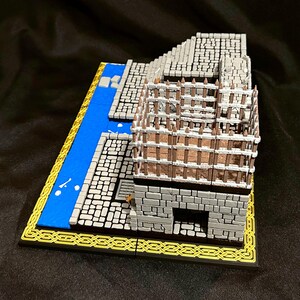 Puede incluir: Modelo en miniatura de un edificio de piedra con una valla de madera, un r&iacute;o azul y un borde amarillo. El edificio es gris con un patr&oacute;n de ladrillos detallado. El r&iacute;o es azul con detalles blancos. La base tiene un borde decorativo negro y amarillo.