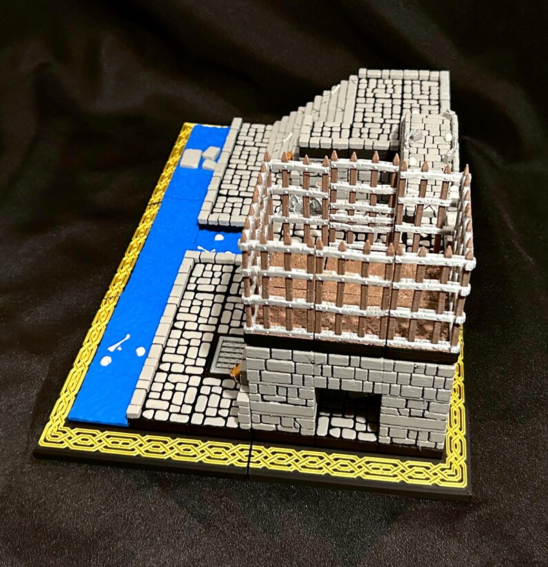 Puede incluir: Modelo en miniatura de un edificio de piedra con una valla de madera, un r&iacute;o azul y un borde amarillo. El edificio es gris con un patr&oacute;n de ladrillos detallado. El r&iacute;o es azul con detalles blancos. La base tiene un borde decorativo negro y amarillo.