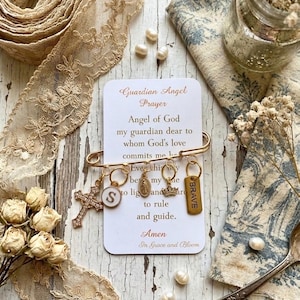 May include: A gold-colored safety pin with attached charms, including a cross, a circular pendant with the letter "S", a Virgin Mary pendant, a crown, and a tag that says "BRAVE". A card with the "Guardian Angel Prayer" is attached.