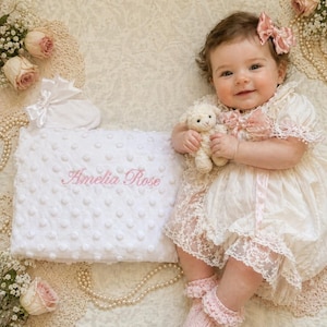 Puede incluir: Un bebé con un vestido de encaje color crema y un lazo rosa. Una manta blanca bordada con "Amelia Rose" y un cordero de peluche también están en la imagen. La escena está decorada con perlas y rosas.