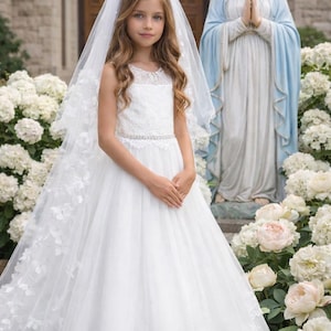 May include: A young girl in a white dress and veil stands in front of a statue and white flowers. The dress features lace detailing and a jeweled belt. The veil is adorned with floral accents.