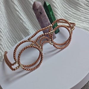 May include: A pair of decorative glasses crafted from twisted copper wire, featuring circular frames and a bridge. Behind the glasses stand two pointed crystal pillars, one in a light purple hue and the other in a deep green, set on a white base.