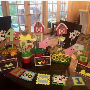 May include: A farm-themed party centerpiece with a red barn, a cow, a pig, and a chicken. The centerpiece is decorated with sunflowers, hay, and a banner that says "I am 1".