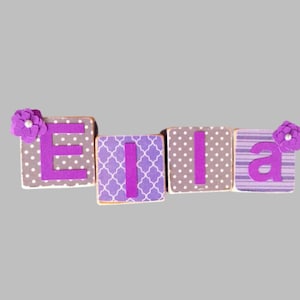 May include: Wooden blocks spelling the name "Ella". The blocks feature the letters in purple, with decorative patterns including polka dots, stripes, and a trellis design. Each end block has a purple flower attached.