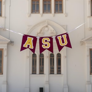 May include: A maroon banner with gold lettering spelling "ASU" hangs in front of a white building with multiple windows. The banner is strung across a white string. The letters have a white outline.