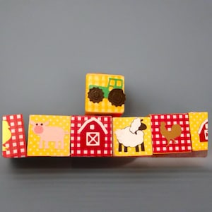 May include: A set of colorful farm-themed blocks. Each block features a different farm animal or object, including a pig, sheep, rooster, and tractor. The blocks have a yellow background with red gingham borders, creating a playful and educational toy.