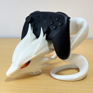 May include: A white dragon-shaped gaming controller stand holding a black controller. The dragon has red and yellow eyes and sharp, pointed details. The stand is designed to cradle the controller securely.
