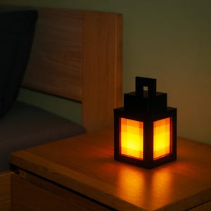 Puede incluir: Una lámpara de noche negra de estilo pixelado que emite un brillo naranja cálido. La lámpara cúbica, con asa, está sobre una mesita de noche de madera. La luz está encendida, iluminando la habitación con una luz suave y ambiental.