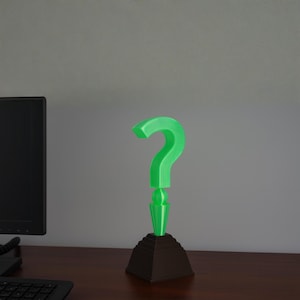 May include: A green question mark sculpture atop a brown stepped base. The question mark is a bright, vibrant green, and the base is a dark brown. The sculpture is likely a decorative item or a desk accessory.