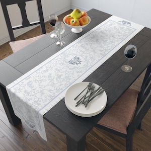 May include: A dining table set for a meal, featuring a white and blue floral patterned table runner. The table is set with plates, forks, wine glasses, and a bowl of apples. The table and chairs are dark wood.