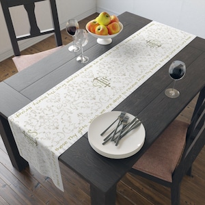 May include: A dining table set for a meal, featuring a white table runner with a floral pattern and text. The table is set with plates, forks, wine glasses, and a bowl of apples. The table is dark wood.