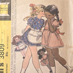 May include: Vintage McCall's 3809 sewing pattern for a child's dress, apron, and doll. The illustration shows two girls in dresses, one in blue floral and the other in red gingham, with matching dolls. The pattern is marked "Carefree Pattern".