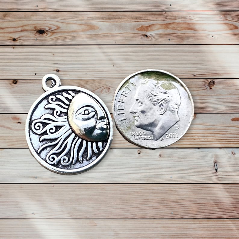 May include: A silver-colored sun and moon charm with intricate details, next to a U.S. dime coin. The charm features a smiling moon face and sun rays. The dime shows the profile of a man and the year 2017.