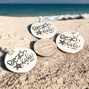 Puede incluir: Tres dijes de plata con el texto "Beach babe" y un peque&ntilde;o dise&ntilde;o de estrella de mar. Los dijes est&aacute;n sobre una superficie arenosa.