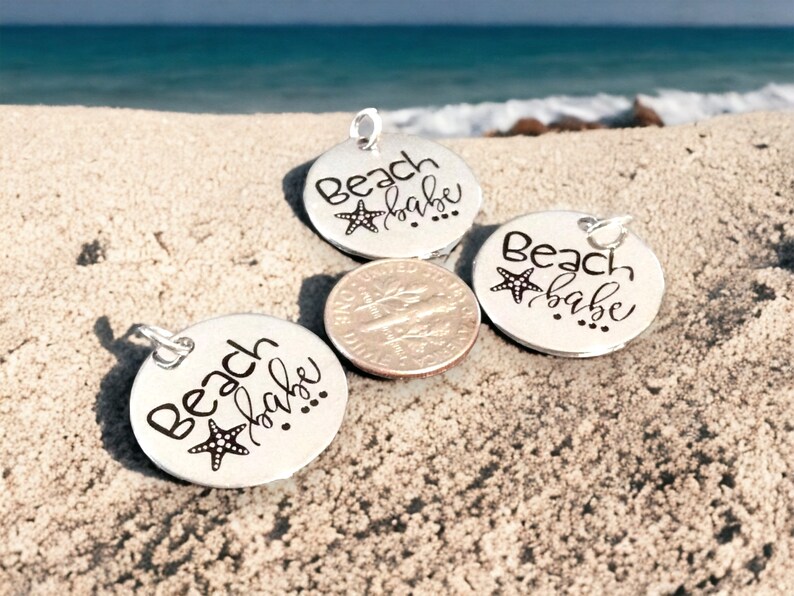 Puede incluir: Tres dijes de plata con el texto "Beach babe" y un peque&ntilde;o dise&ntilde;o de estrella de mar. Los dijes est&aacute;n sobre una superficie arenosa.