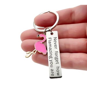 May include: A silver keychain with a pink flamingo charm and a rectangular tag. The tag is engraved with the words "Never forget how Flamazing you are". The flamingo charm has gold legs and a pink body.