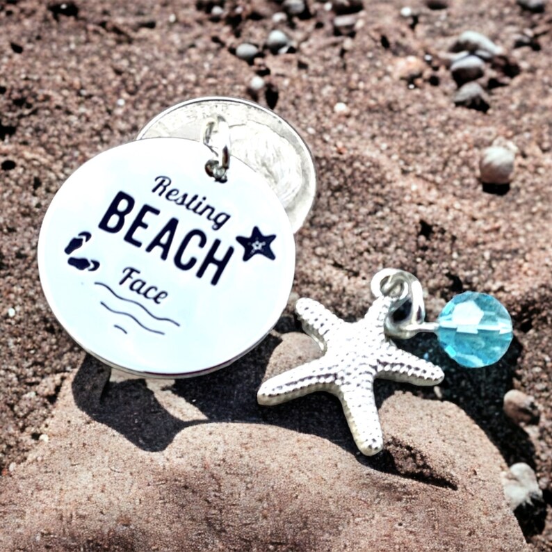 May include: A silver charm with the text "Resting Beach Face" and a small blue crystal charm attached to a silver starfish.