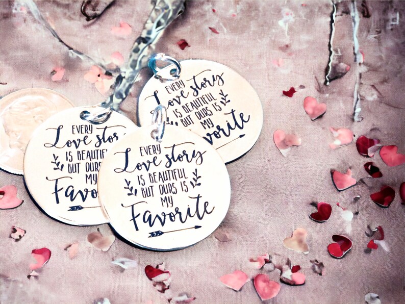 May include: Three round wooden signs with the text "Every love story is beautiful but ours is my favorite" on each sign. The signs are on a pink surface with pink and red confetti hearts.
