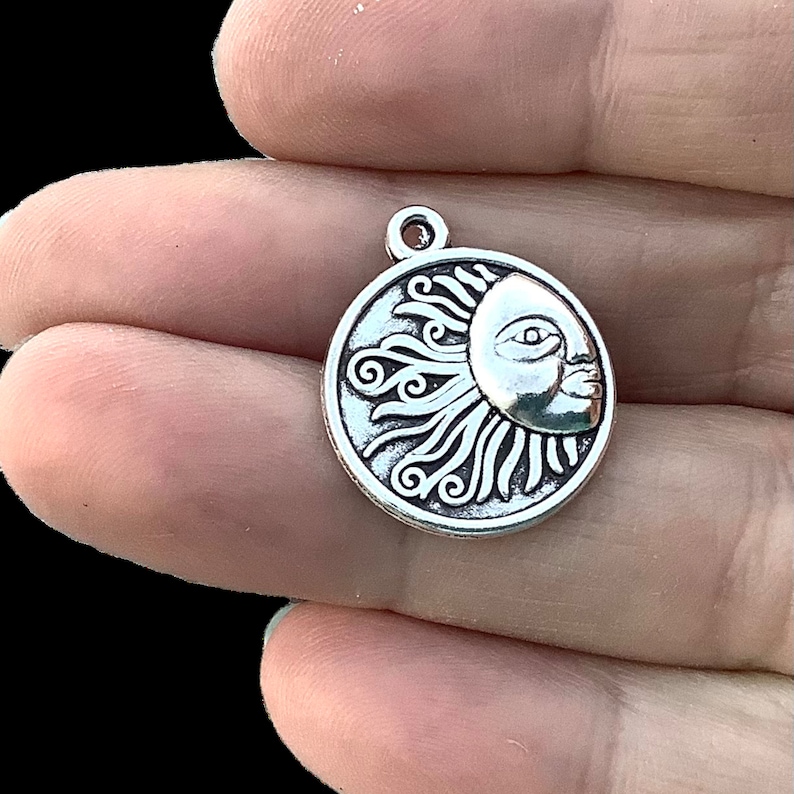 May include: A silver-colored charm featuring a sun face design. The sun has a detailed face with closed eyes and flowing rays. The charm is round with a small loop at the top for attaching to jewelry or other crafts.