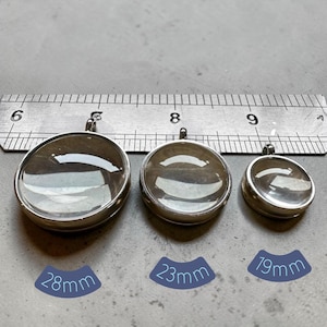 May include: Three round silver-toned metal pendant settings with clear, domed glass cabochons. A metal ruler is positioned above the pendants, and labels indicate the diameters as 28mm, 23mm, and 19mm for each respective pendant size.