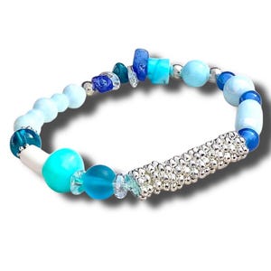 May include: A beaded bracelet featuring a mix of blue and turquoise beads, with a silver-colored cylindrical accent. The bracelet includes various shapes and sizes of beads, creating a unique and eye-catching design. The bracelet is designed to fit a standard wrist size.