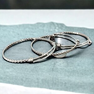 May include: Three silver bracelets, one with a braided design and two with a simple, smooth design. The bracelets are stacked on top of each other.
