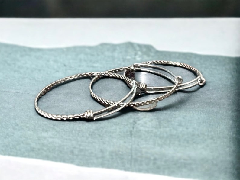 May include: Three silver bracelets, one with a braided design and two with a simple, smooth design. The bracelets are stacked on top of each other.