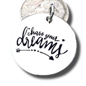 May include: A round, white pendant with the phrase "Chase your dreams" in black script. The design includes an arrow and stars. A silver-colored metal loop is attached at the top.