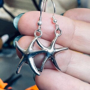 May include: A pair of silver starfish earrings. Each earring features a starfish charm suspended from a silver hook. The starfish have five arms and are detailed with textured surfaces. The earrings are designed for pierced ears.