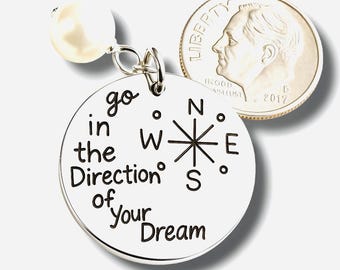 Puede incluir: Colgante plateado con el texto "go in the Direction of your Dream" y un diseño de rosa de los vientos. Una perla blanca cuelga de la parte superior. Una moneda de diez centavos estadounidense está al lado.