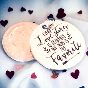 May include: A round silver charm with the text "Every love story is beautiful but ours is my favorite" engraved on it. The charm is surrounded by small red and blue hearts.