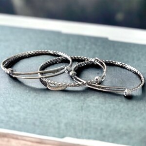 May include: Three silver bracelets, two are thin and one is thicker and braided. The bracelets are stacked on top of each other.