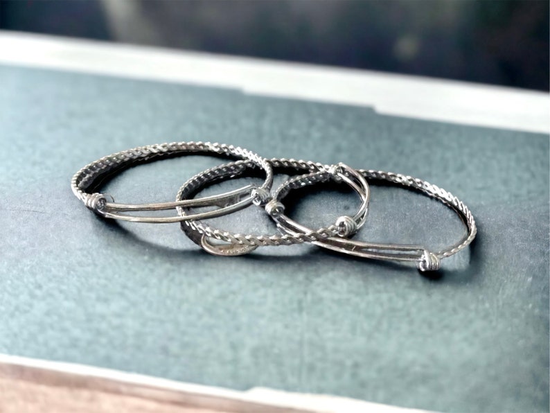 May include: Three silver bracelets, two are thin and one is thicker and braided. The bracelets are stacked on top of each other.